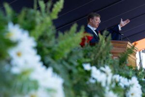 Photo of a man in glasses standing at a podium and clapping. He is wearing red and navy blue academic robes. In the foreground in front of him are white flowers with green foliage.