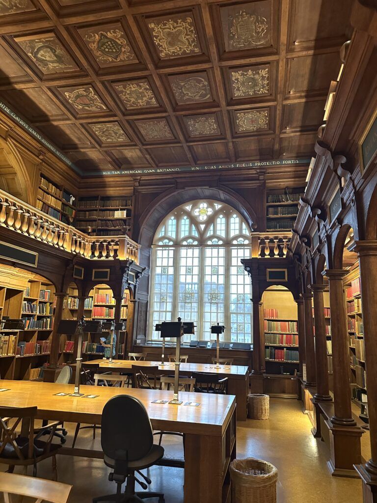 Photo of a wood-paneled library reading room with inset ceiling and a large arcaded window at one end. A number of long tables with reading lamps fill the floor of the room.
