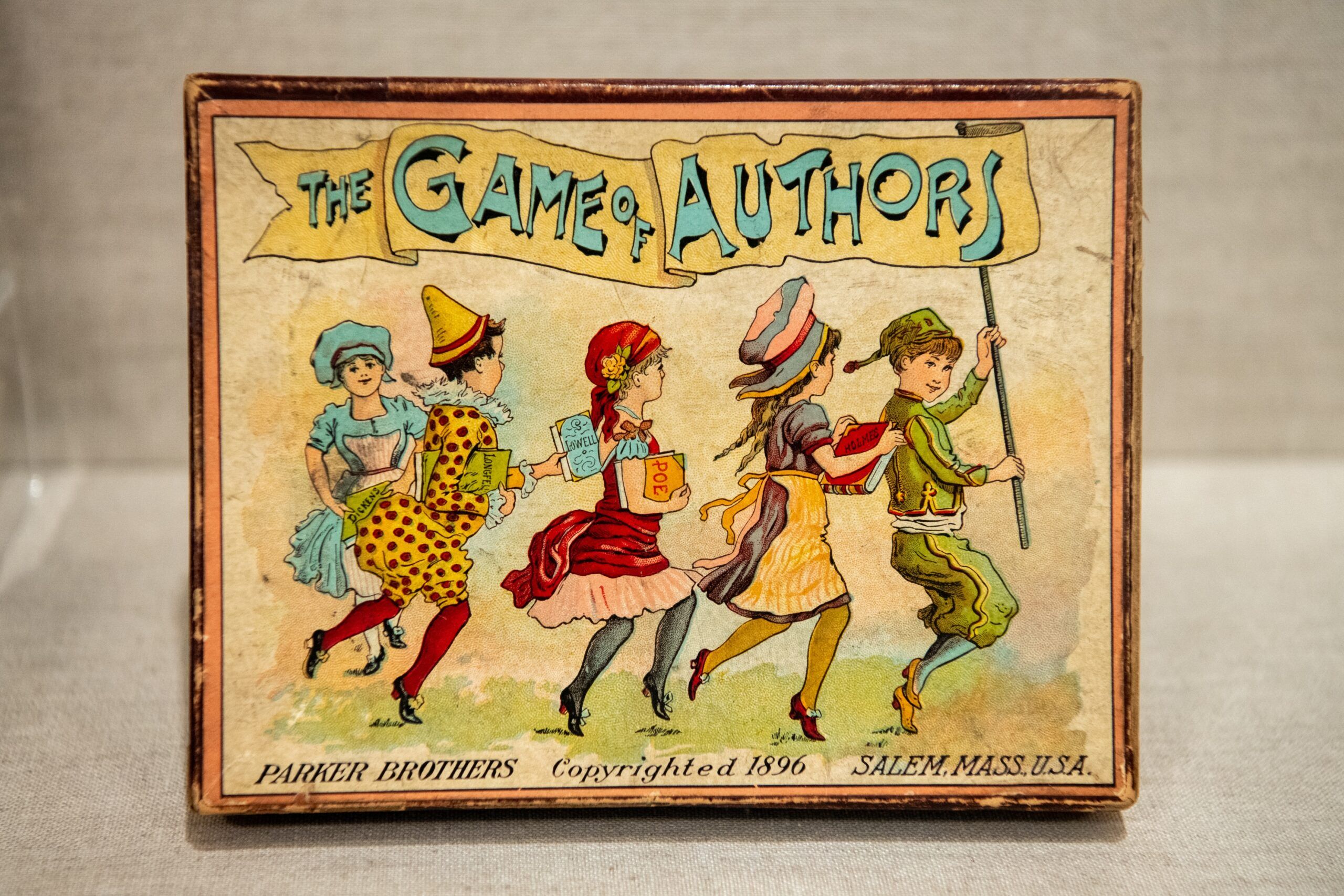 The front of an old card game box featuring several children in colorful costume skipping in a line while the front boy holds a banner reading "The Game of Authors"