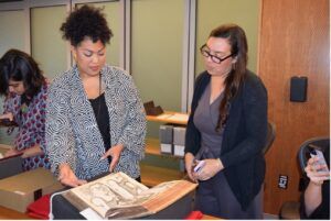 A photo of two women standing behind a table on which are several books. They are examining a large-format book lying open before them.