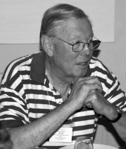 Photo of a man with glasses wearing a striped shirt with a collar. His elbows are resting on a table and he has his hands clasped as he looks off camera.