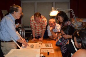 Photo of a group of people gathered closely around a table on which are several old documents. A man on the left points at one of these while the others lean in to view it.