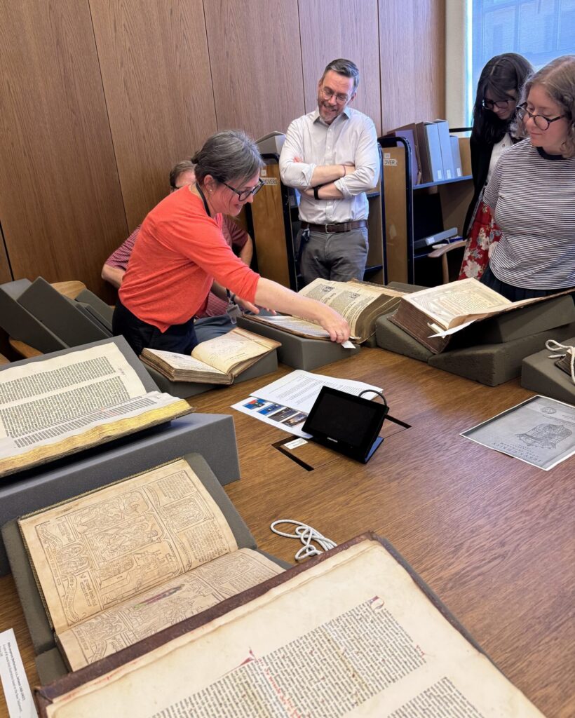 Photo of a woman leaning over a table covered with old books and manuscripts as she points out features to several other people gatherer around the table.