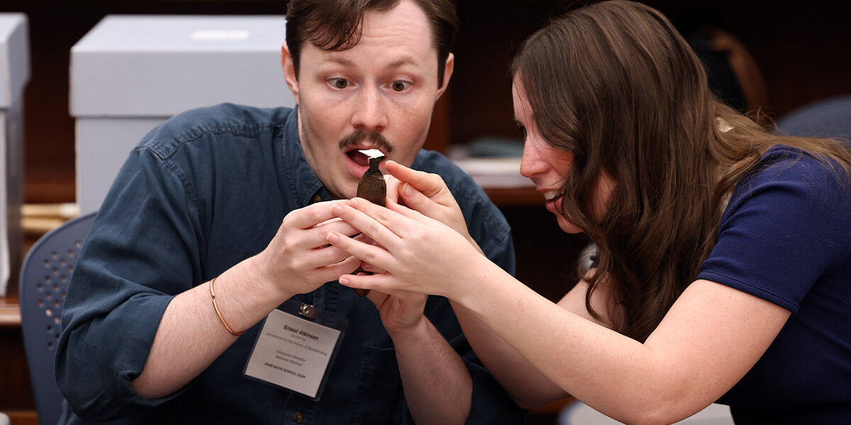 Two Rare Book School students closely examine a leather bookbinding tool together, reacting with curiosity as they study the small hand tool.