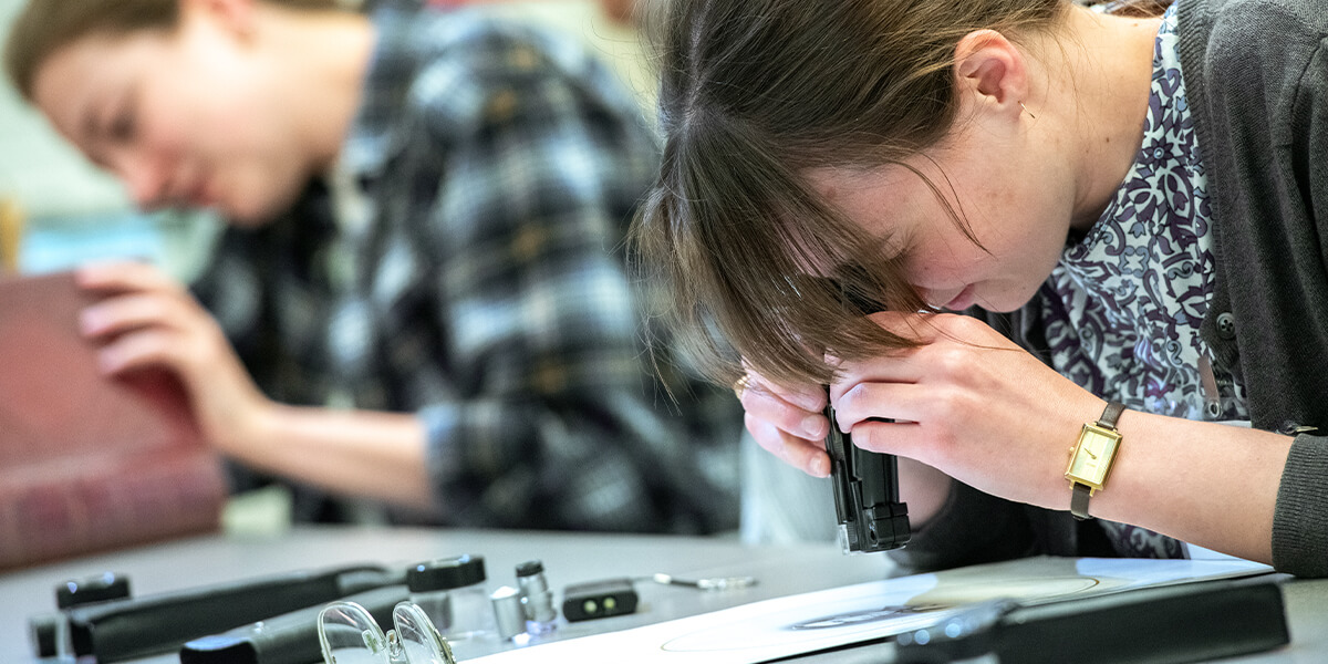 Student closely examines a printed page with a handheld magnifier while another student studies a book in the background at Rare Book School.