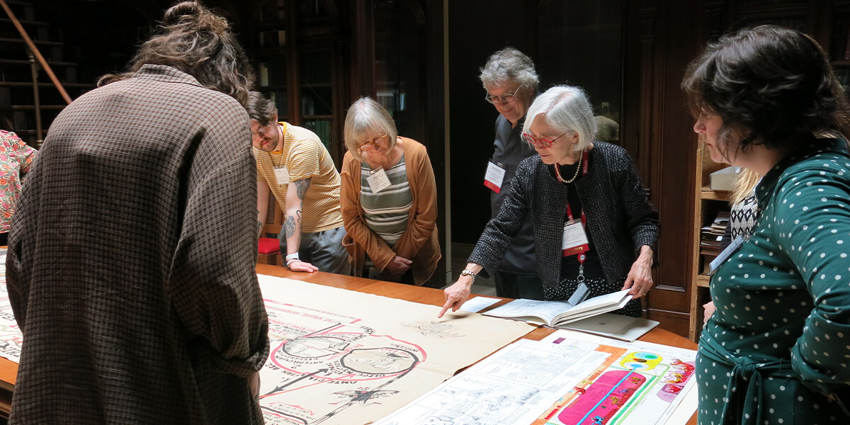 Instructor points to a large historic print on a table as Rare Book School students gather around to examine it.