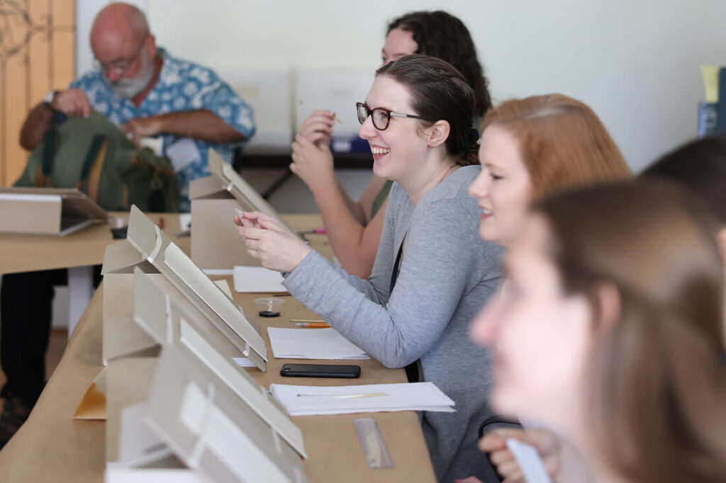 Students in a Rare Books School class sit at angled writing desks, practicing with quill pens and ink while smiling and engaging with the lesson.