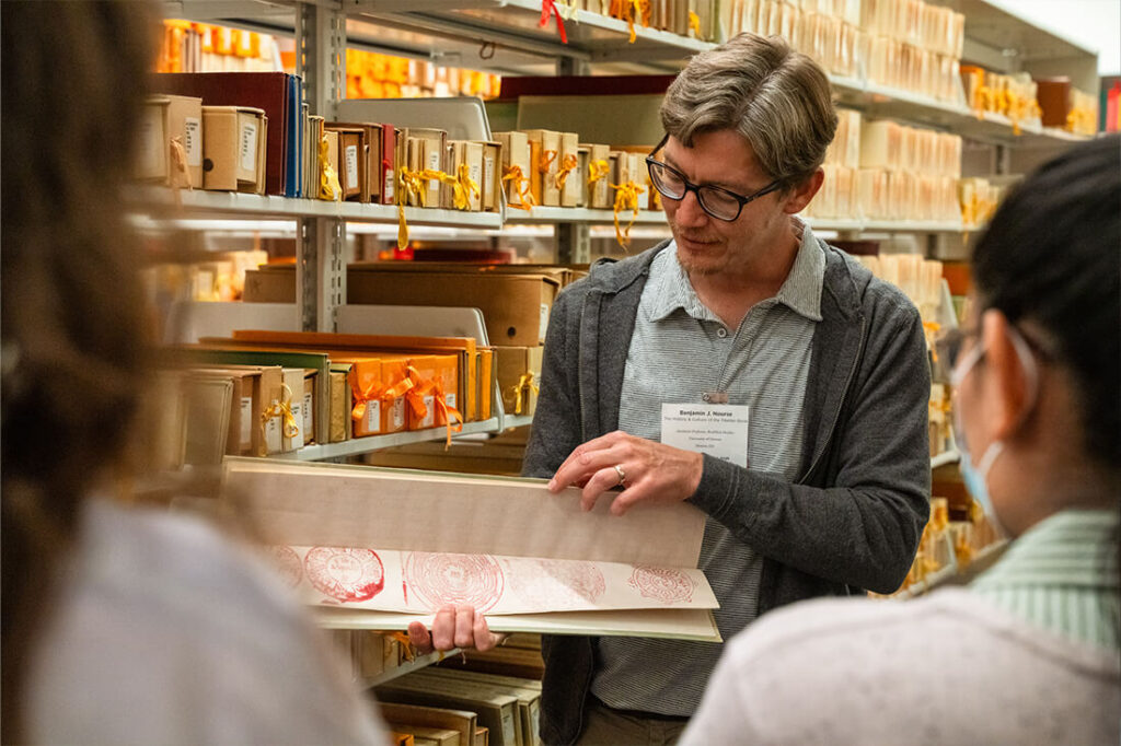 Instructor at Rare Book School holds up a sheet with red ink impressions, pointing out details to students during a demonstration in a library stacks area.