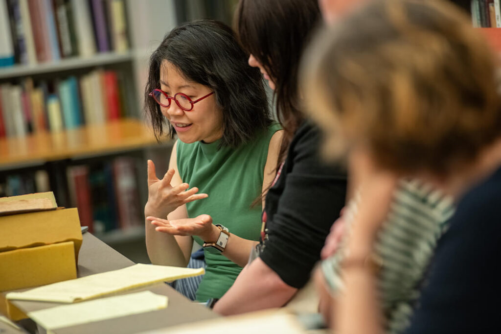 Students sitting at table, surrounded by shelves of books, having a discussion with rare books and note pads in front of them.