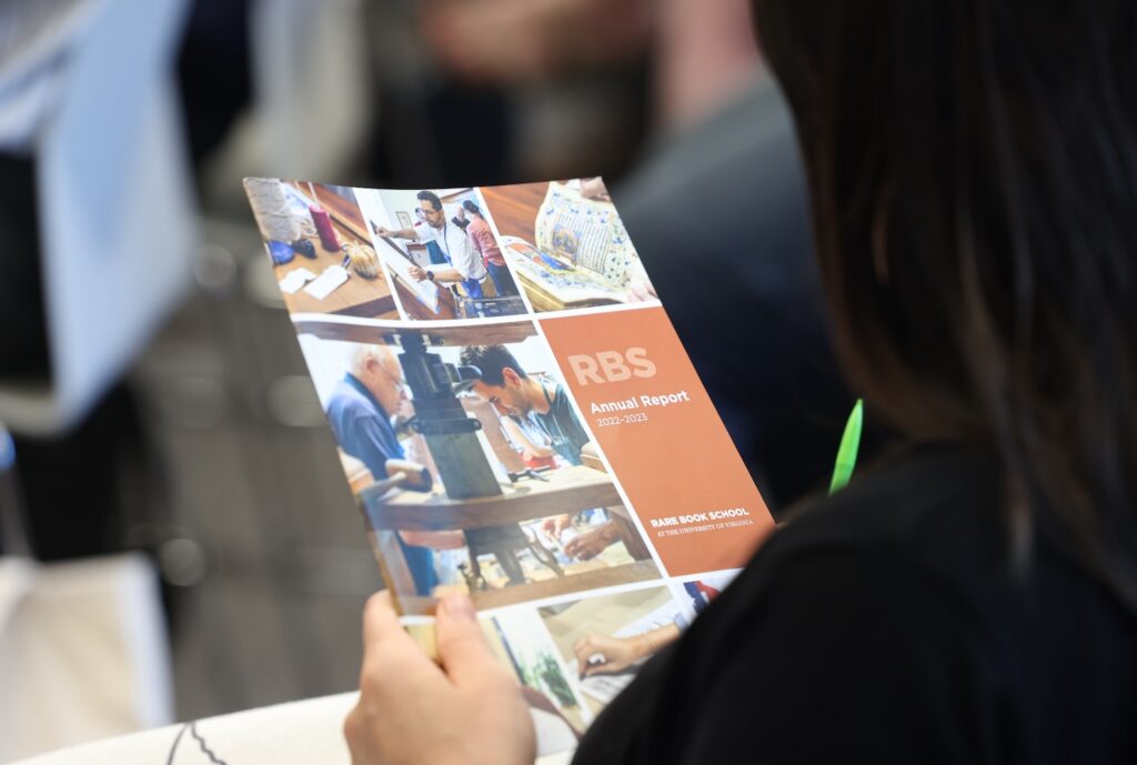 A person with dark hair holds up a copy of the 2022–2024 RBS Annual Report, which has a cover showing blocks of photos of people with rare books and a printing press, alongside a block of orangey-red where the report's title is.