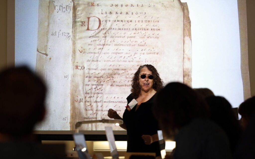 Photo of a woman teaching in front of a projection screen in a darkened room. On the screen is displayed a page of a medieval manuscript.