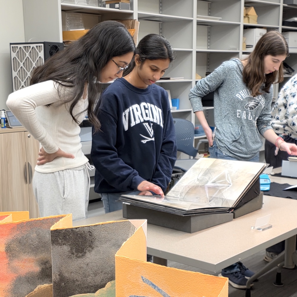Two young women, one of them in a shirt that says "Virginia" with the UVA logo below it, look at a modern book with metal pages. Beside them another young woman looks at another book, and in the foreground are folded out accordion-style pages of a book made with bright colored paper.