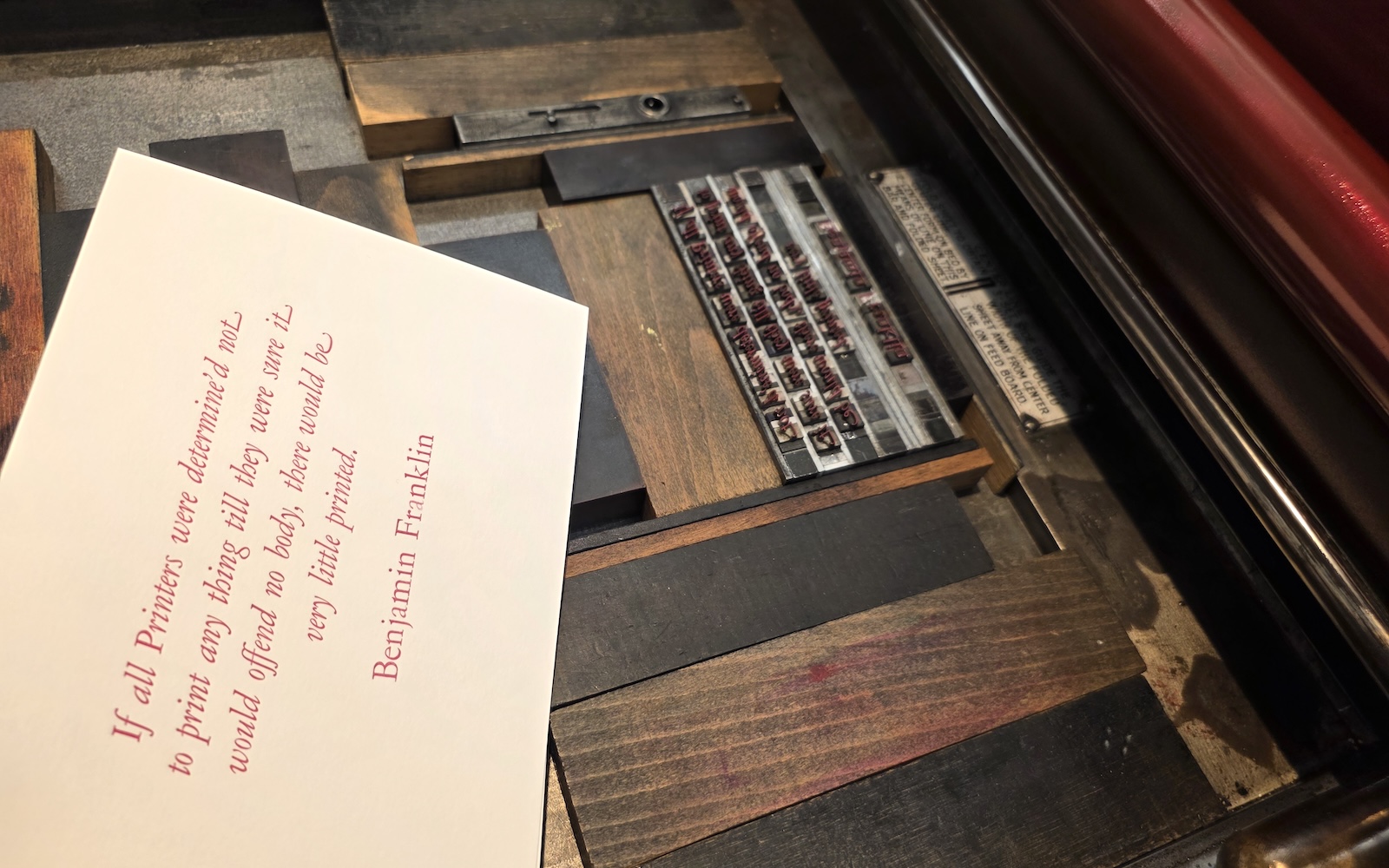 A printed notecard rests on the blocks of a letterpress with red ink on its rollers and several lines of set text. The card reads, in red ink, "If all Printers were determine'd not to print any thing till they were sure it would offend no body, there would be very little printed. Benjamin Franklin".