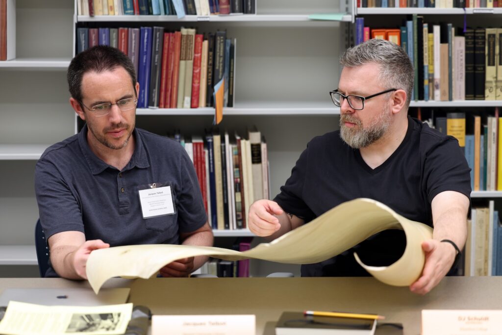 Two men sit at a table examining a long sheet of parchment that is rolled out between them