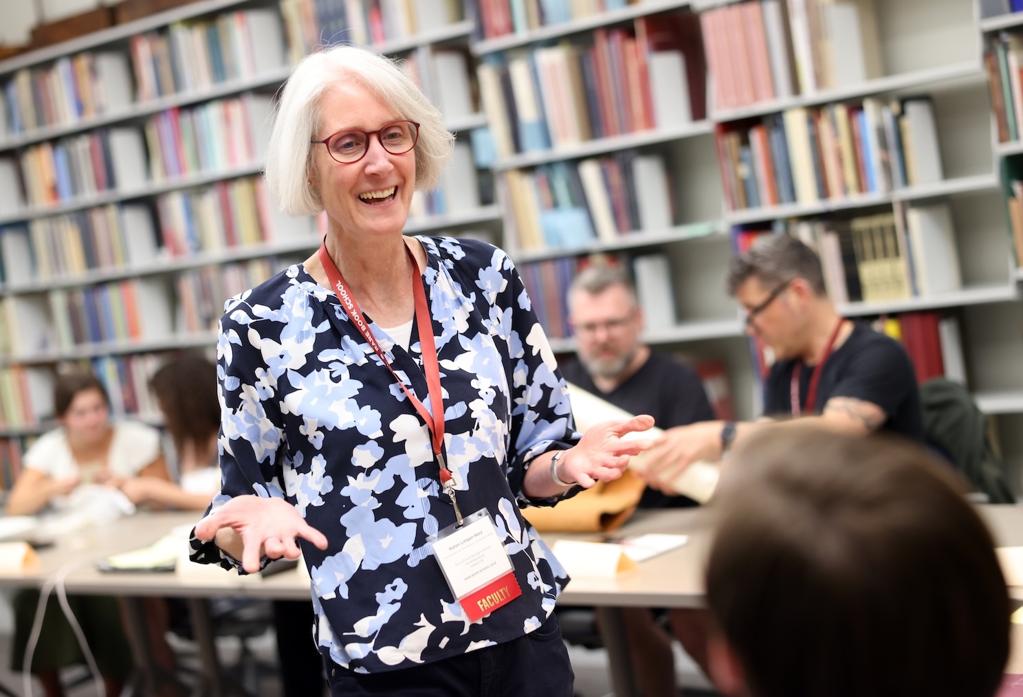 A smiling woman wearing a Rare Book School faculty name badge holds her hands out as she speaks to students seated around a table in a bookshelf-lined room.