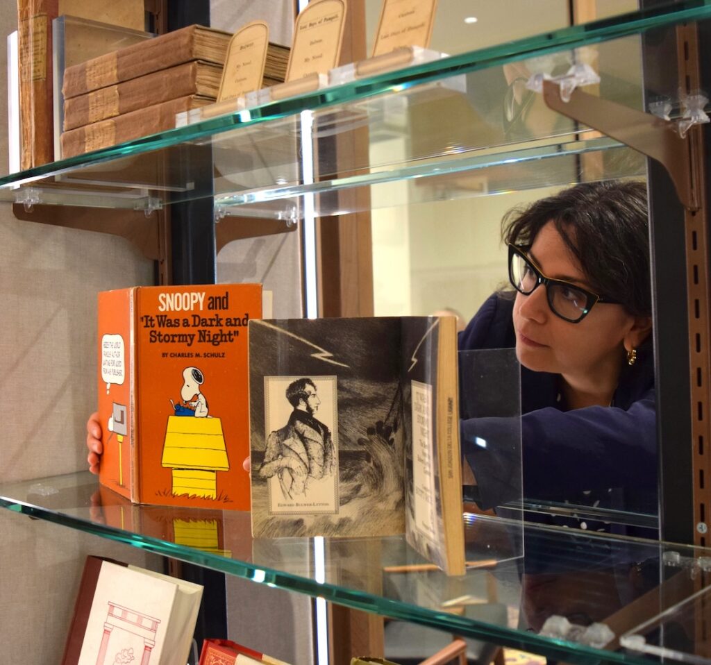 Photo of a dark-haired woman with glasses leaning over to adjust a book on display in a glass-shelved display case.