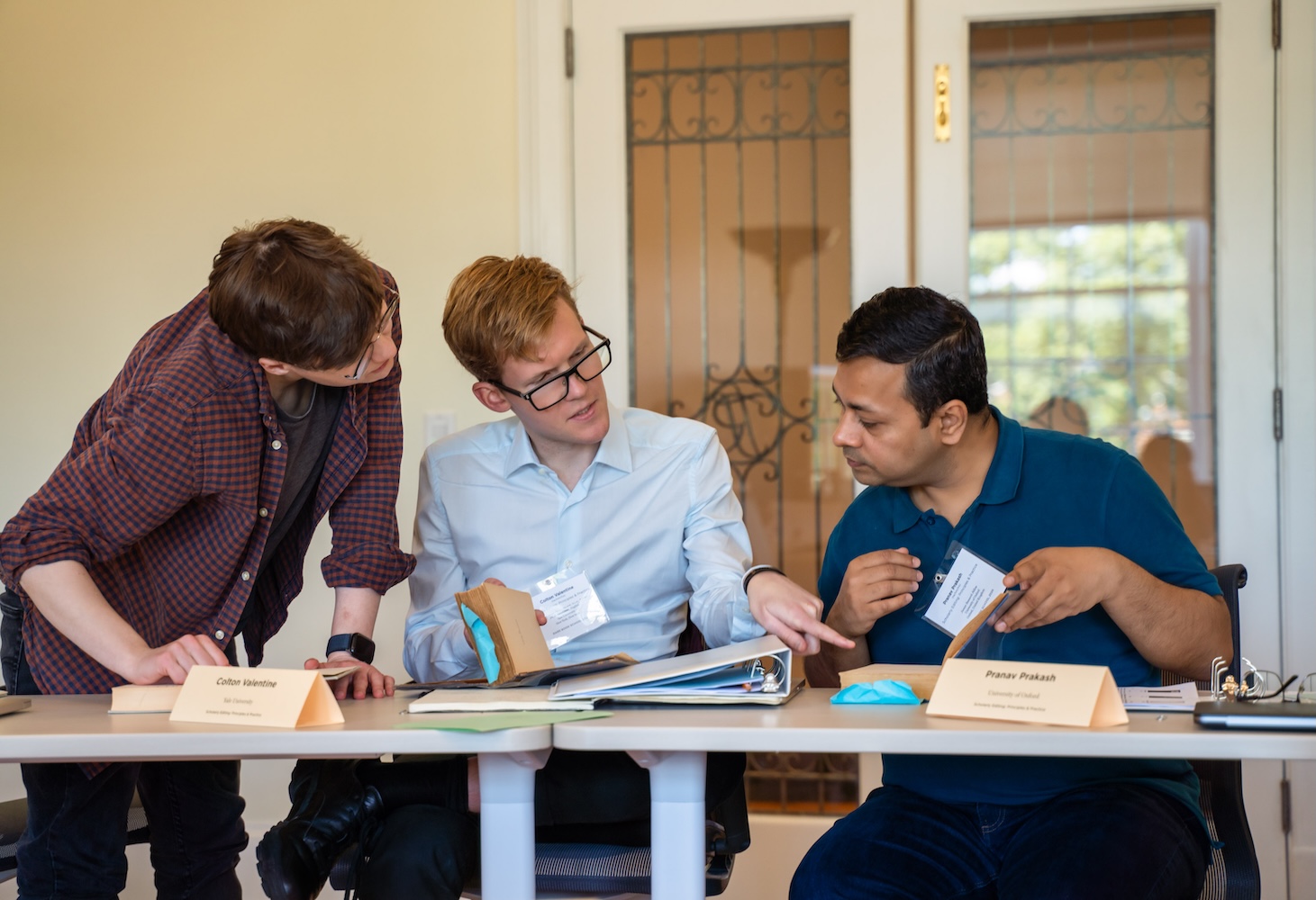 A group of three men are gathered around a long table, looking at a couple of rare books laid out upon it.