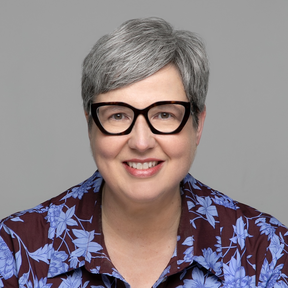 Headshot of a smiling woman with short gray hair and glasses, wearing a burgundy shirt with lavender flowers on it.