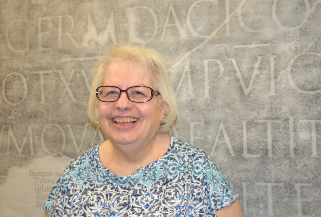 Photo of a woman with glasses smiling at the camera in front of a rubbing of a Latin monumental inscription