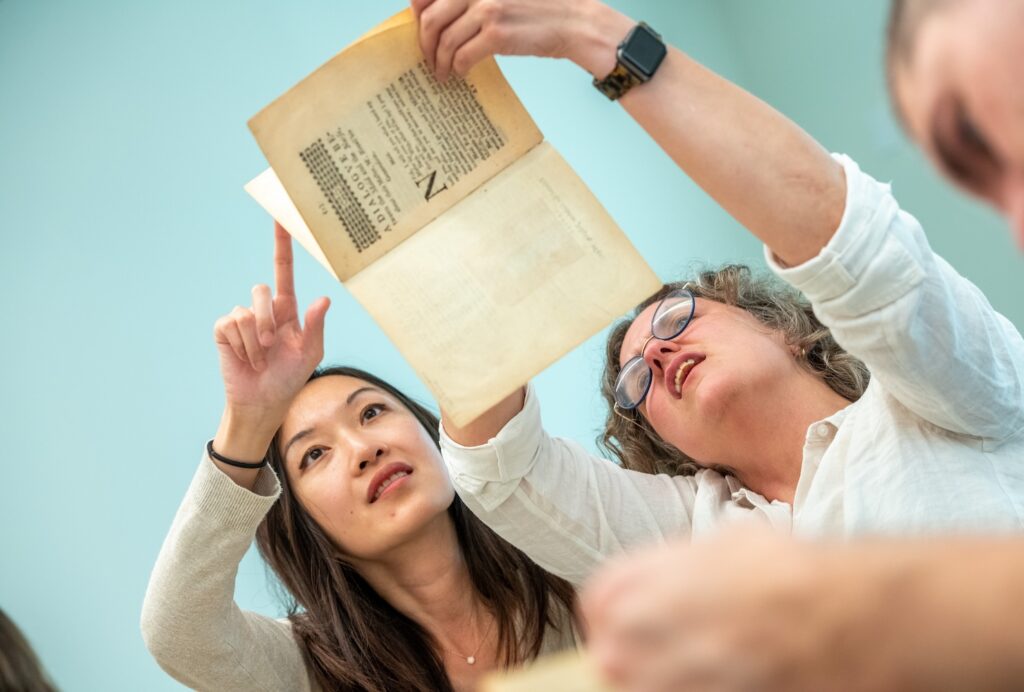 Two women hold a book up over their heads as they examine details of the pages. The wall behind them is turquise.