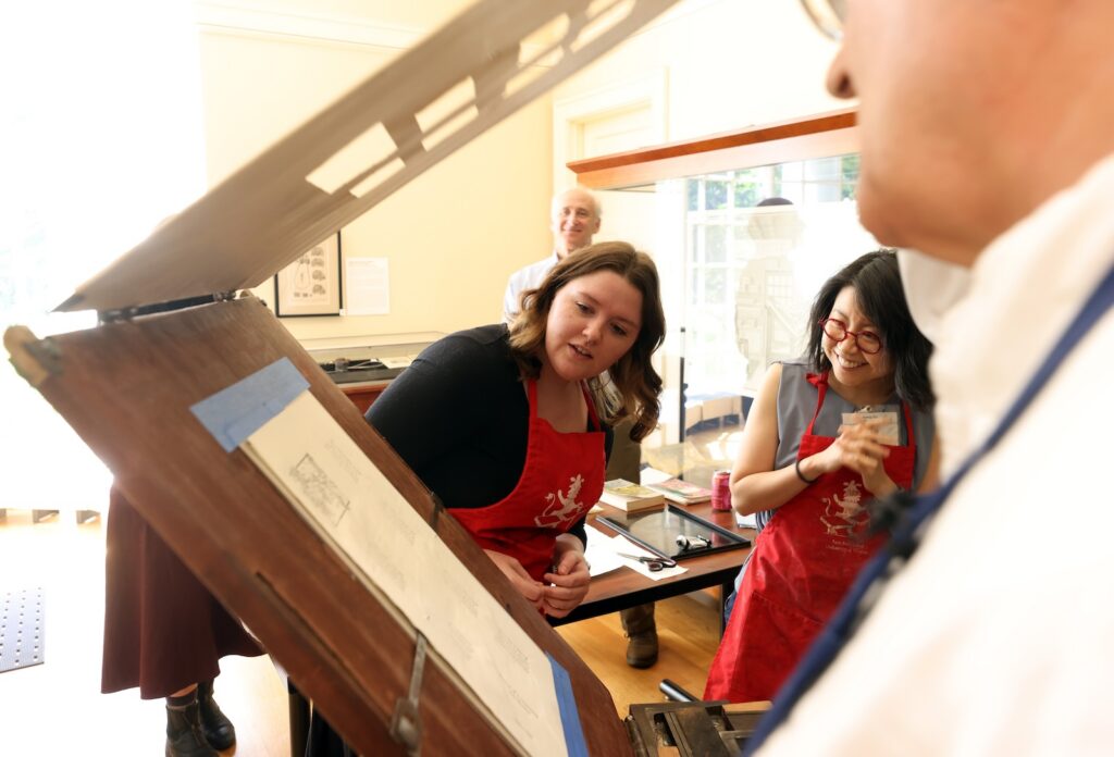 Two women in red aprons examine a print made on a large hand press