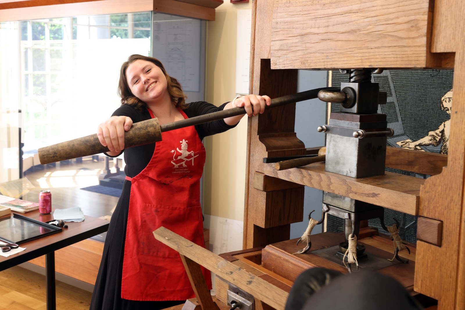 Photo of a woman in a black dress and red apron operating a wooden hand press