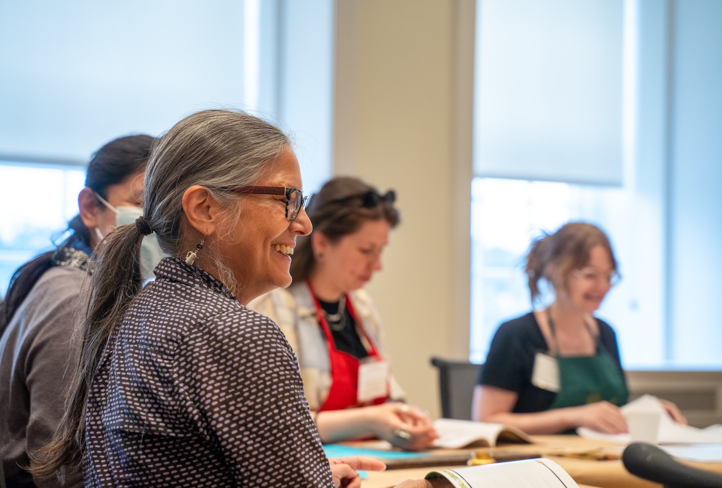 Several women sit around a table. Various notebooks and other items are on laid out on the table top.