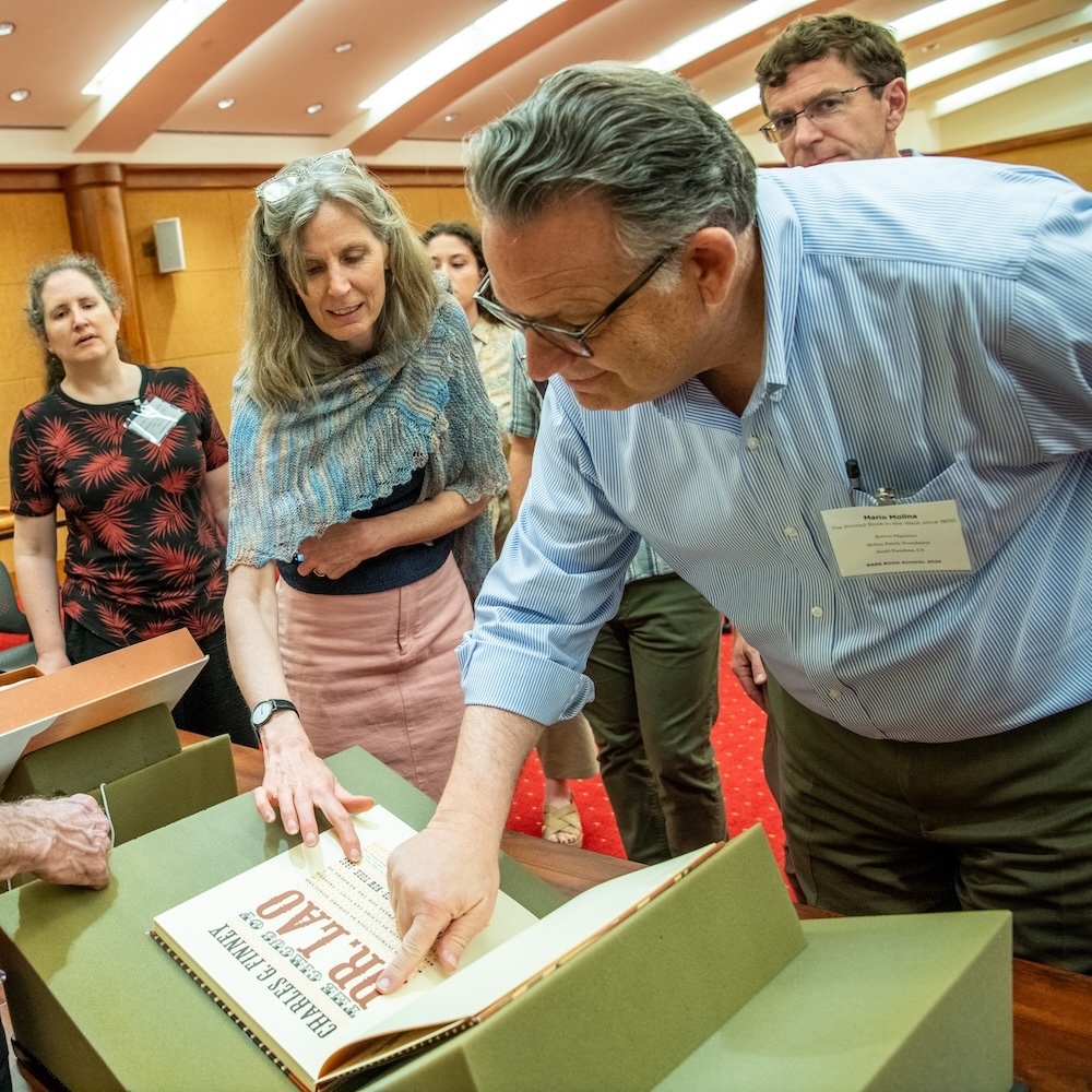 A group of RBS students gather around a rare book that is laid out on foam rests on a table. Their faculty member looks on from the other side of the table.
