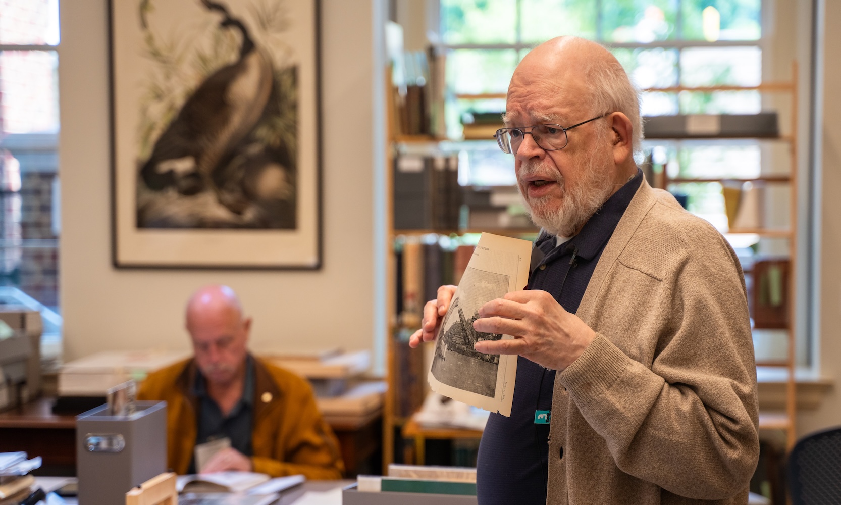 Photo of a man in a tan cardigan and glasses holding up an old document to people off camera.