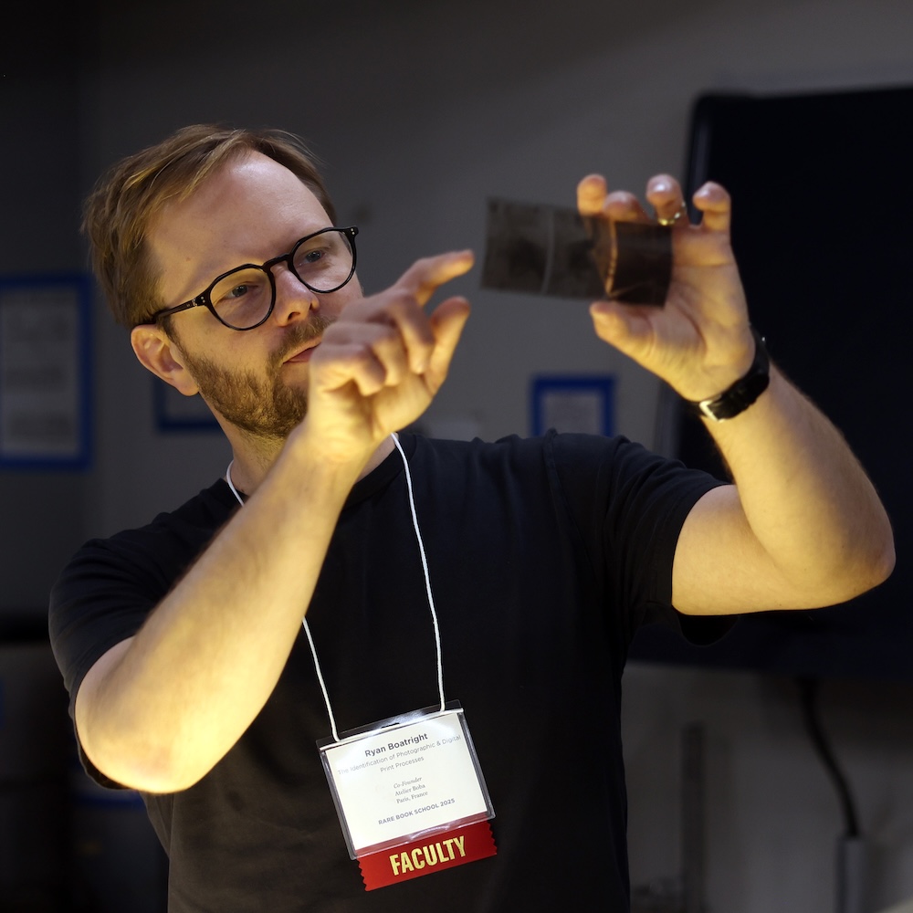 Photo of a man in a black shirt standing in a dark room holding up some photo negatives and pointing to a detail on them. He has an RBS faculty lanyard around his neck.
