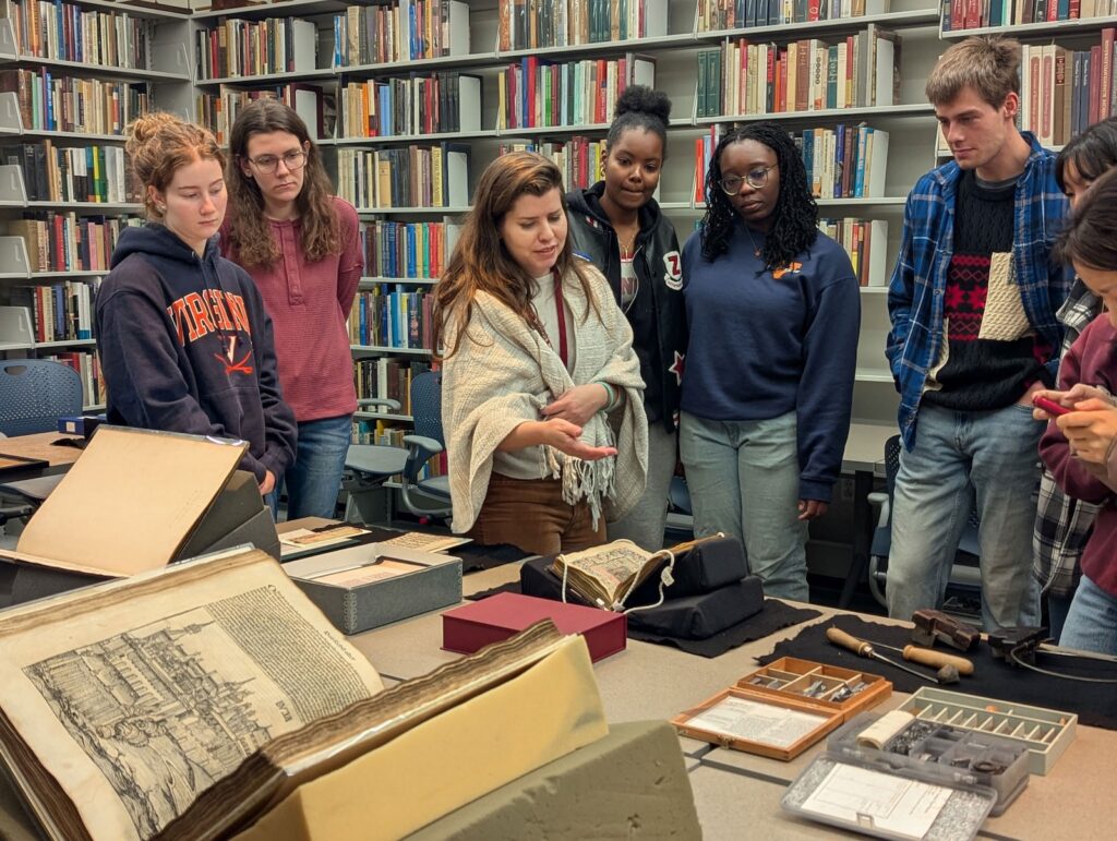 A woman in a shawl points out details of a rare book to a group of students gathered around tables on which a number of rare books are laid out on display. The wall behind them in lined with books on shelves.