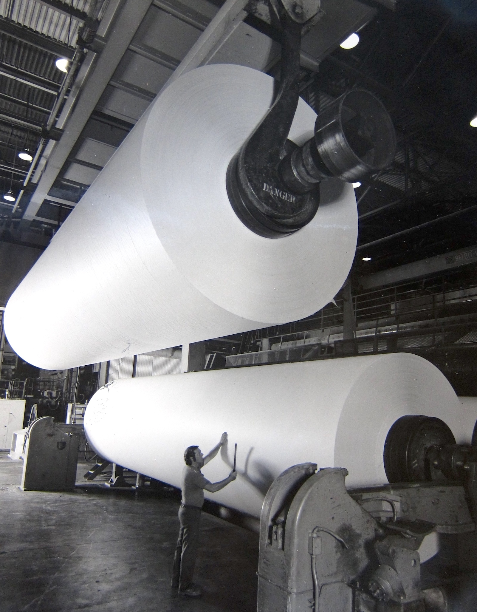 Old black and white photo of a man examining giant rolls of newsprint held up by mechanical lifts