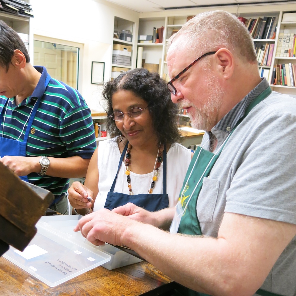 A woman in a blue apron and a man in a green apron, both wearing glasses, work on setting type