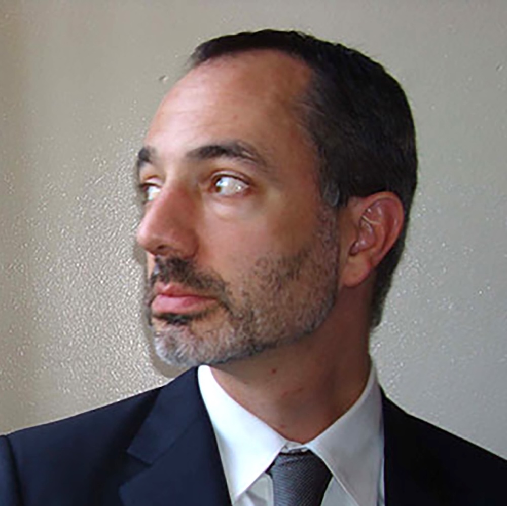 Headshot of a dark-haired man in a suit with his head turned to look to his right (viewer's left)