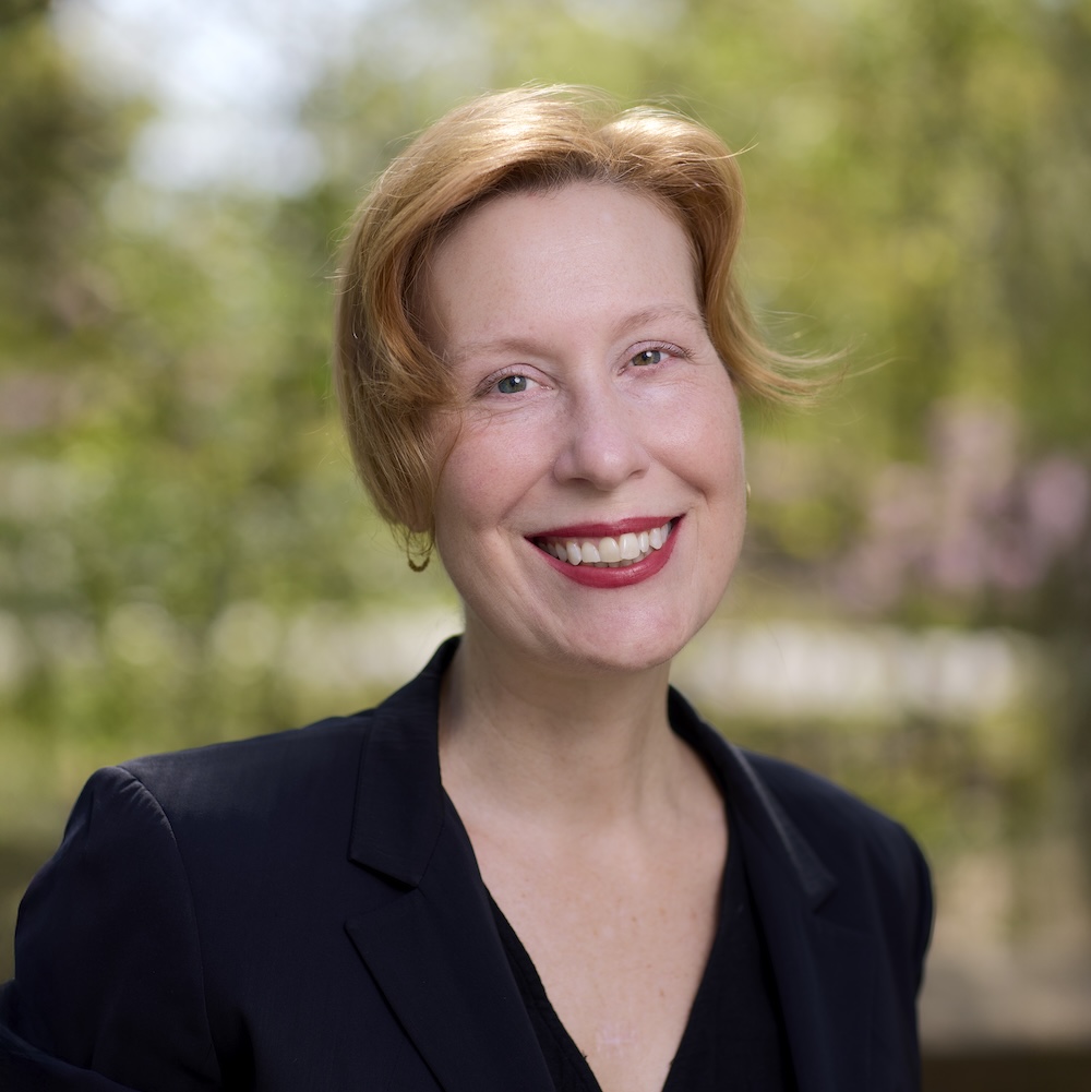 Headshot of a smiling woman in a V-necked top standing against a blurred background of trees