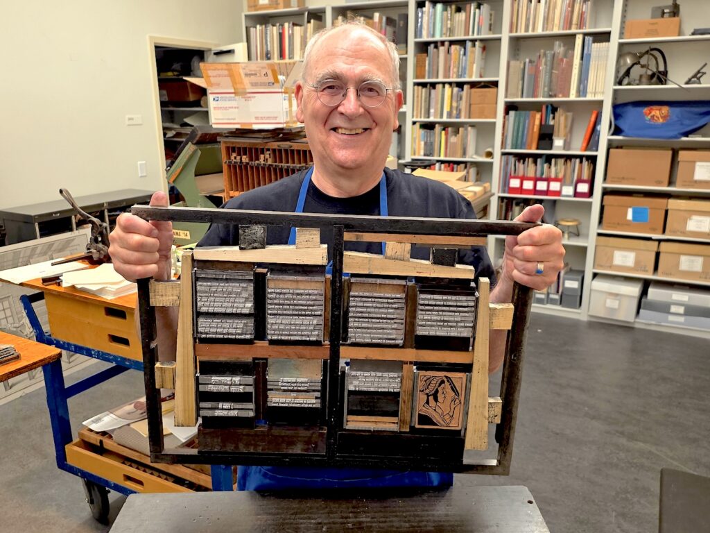 A man in glasses and a blue apron stands in the bookshelf-lined RBS Print Office. He is is smiling as he holds up a block of locked-in type, which also includes a woodcut of poet Phillis Weatley in a bonnet holding a quill pen.