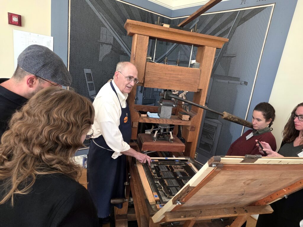 A man in a white shirt and blue apron stands alongside the open bed of a large wooden hand press as he explains the press to a group of RBS students gathered around him.