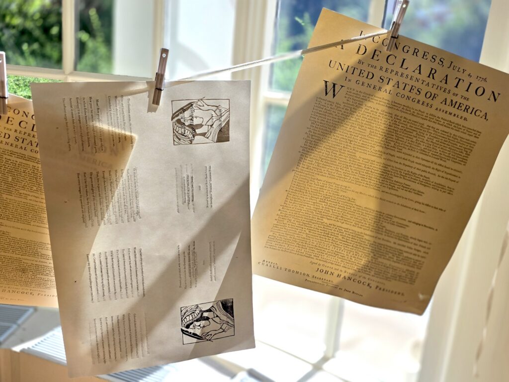 Several sheets of printed paper hang by pins from a line strung in front of a large window. Two of the visible pages are broadside copies of the Declaration of Independence, while the sheet in the middle has a woodblock image of Phillis Wheatley alongside her poetry.