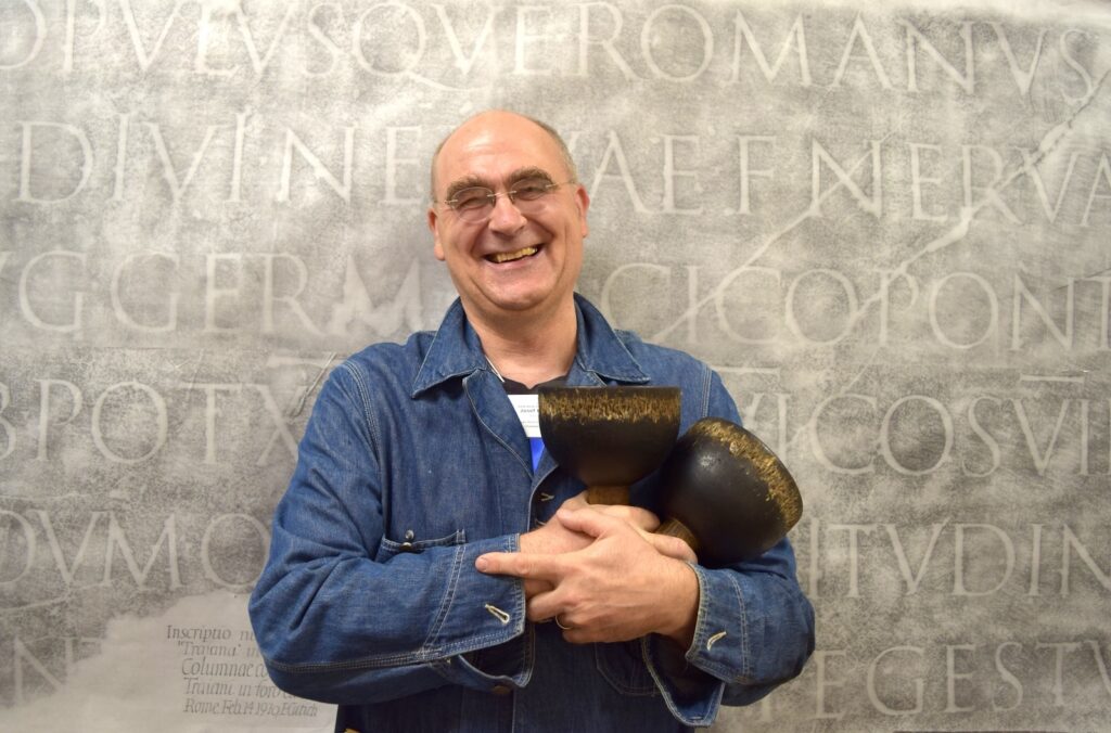 A man in a blue denim shirt with a nametag around his neck smiles at the camera as he holds up two wood-handled ink balls. Behind him as a backdrop is a rubbing of a Latin inscription from Trajan's Column.