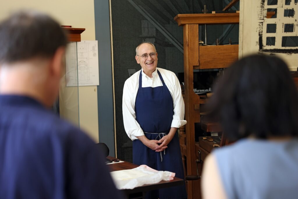 A laughing man in glasses, a white button-up shirt, and a dark blue apron stands next to a large wooden hand press. Opposite him, with their backs to the camera, are a man and a woman with blue shirts.