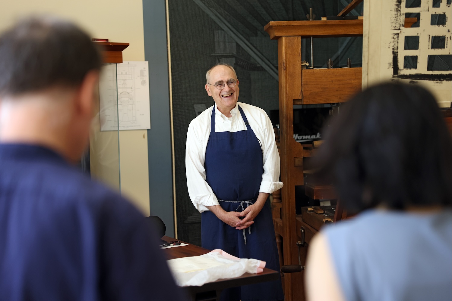 A laughing man in glasses, a white button-up shirt, and a dark blue apron stands next to a large wooden hand press. Opposite him, with their backs to the camera, are a man and a woman with blue shirts.