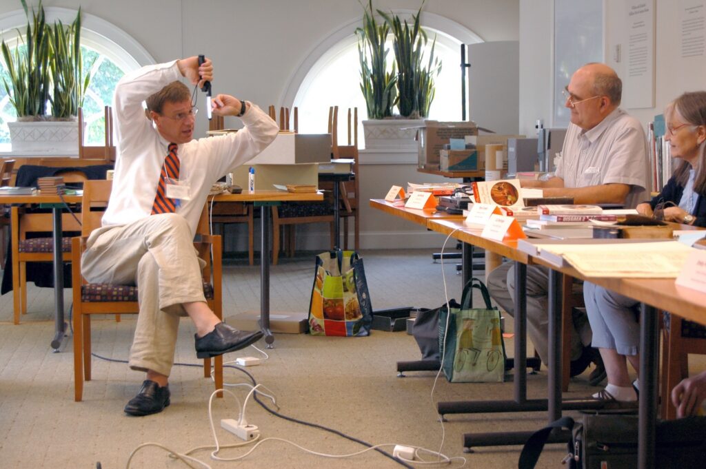 A man in a white shirt, tan pants and a blue-and-orange UVA striped tie holds up a portable LED light to demonstrate its use for students who are seated at a table to one side of him. The table is covered with books and notepads.