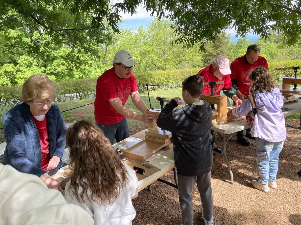 A group of people in matching red shirts with the Declaration of Independence printed on it stand behind a set of trestle tables set up outside under a tree. They are showing three children with their backs to the camera how to operate BookBeetle presses.