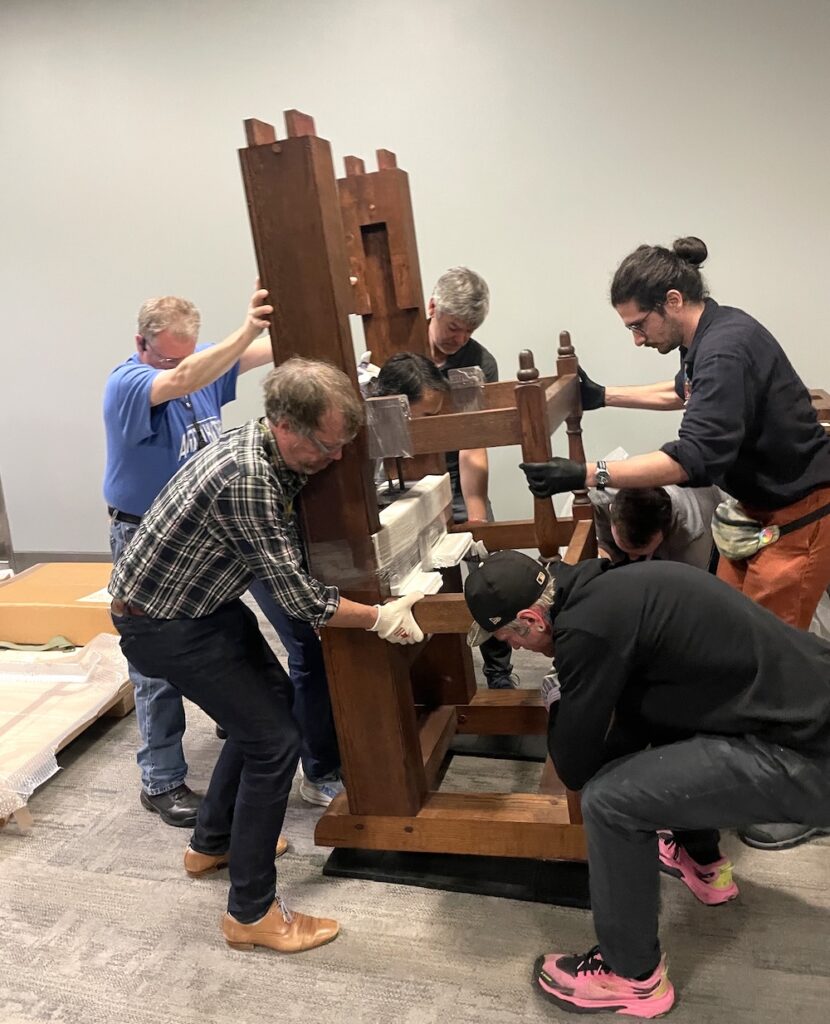 A group of men carefully lowers a large wooden hand press into place on a carpeted floor