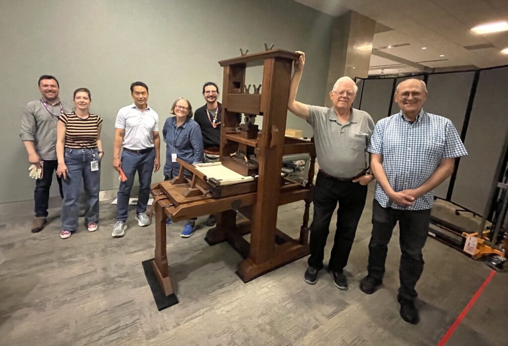 Seven smiling people (two women and five men) stand on either side of a large eighteenth-century style wooden hand press. The press is set up near an unmarked grayish wall.