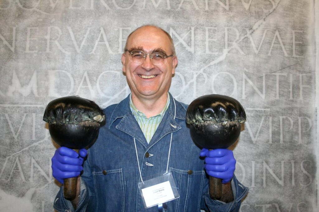 A man in a blue denim shirt with a nametag around his neck smiles at the camera as he holds up two wood-handled ink balls. Behind him as a backdrop is a rubbing of a Latin inscription from Trajan's Column.