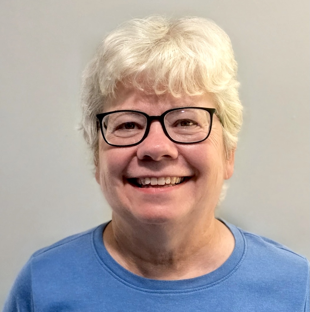 A smiling woman with short light-colored hair, dressed in a blue shirt and wearing black-rimmed glasses
