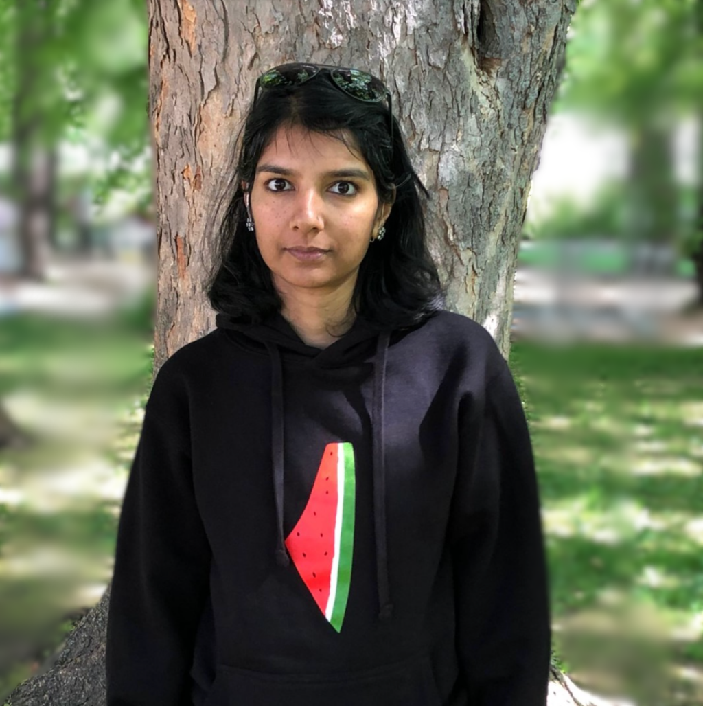 A dark-haired woman in a black hoodie with a picture of a watermelon slice on it stands in front of a tree trunk in tree-filled park.
