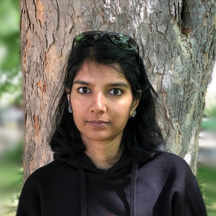 A dark-haired woman in a black hoodie with a picture of a watermelon slice on it stands in front of a tree trunk in tree-filled park.