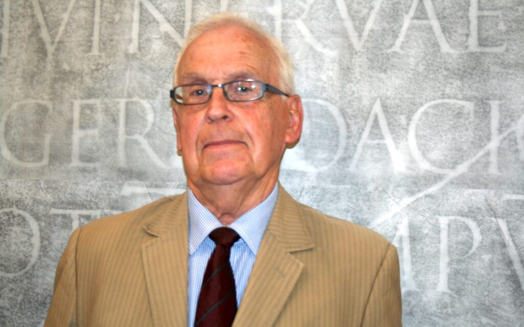 A headshot of a main in glasses wearing a dark tie, a blue-checked collared shirt, and a striped tan suit jacket. Behind him as a backdrop is a rubbing of a Latin inscription from Trajan's Column.